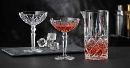 The NACHTMANN Noblesse pitcher filled with a red drink and ice cubes and two NACHTMANN Noblesse cocktail glasses, one of them filled with the same red drink. In the background there is the empty cocktail glass and ice cubes.