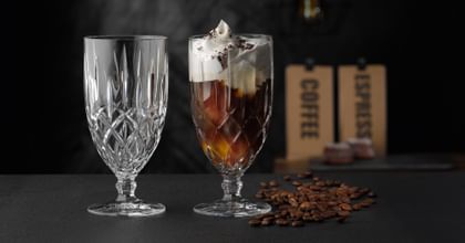The NACHTMANN Noblesse iced beverage glass filled with an iced coffee with cream on it. On the left the same glass but empty. On the right, as a decoration, are coffee beans.
