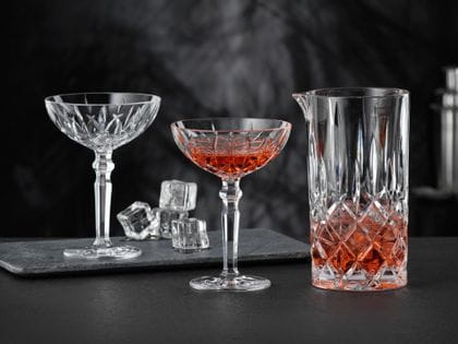 The NACHTMANN Noblesse pitcher filled with a red drink and ice cubes and two NACHTMANN Noblesse cocktail glasses, one of them filled with the same red drink. In the background there is the empty cocktail glass and ice cubes.<br/>