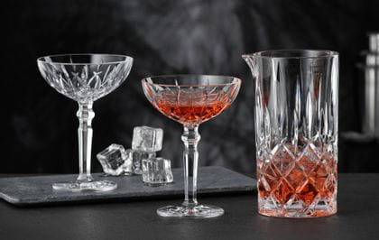 The NACHTMANN Noblesse pitcher filled with a red drink and ice cubes and two NACHTMANN Noblesse cocktail glasses, one of them filled with the same red drink. In the background there is the empty cocktail glass and ice cubes.<br/>