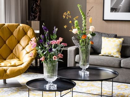 The impressive, with bouquets of flowers filled, NACHTMANN Minverva vases on round side tables in a living room. In the room there are a couch, a lamp, a leather armchair and a carpet with yellow pattern.<br/>