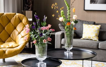 The impressive, with bouquets of flowers filled, NACHTMANN Minverva vases on round side tables in a living room. In the room there are a couch, a lamp, a leather armchair and a carpet with yellow pattern.<br/>