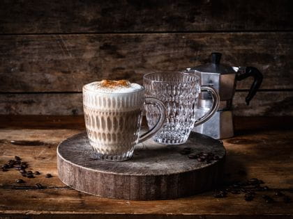 Two NACHTMANN Ethno hot beverage mugs on a bold wooden cutting board. One mug is filled with cappucino, the one behind is empty. In the background is an Italian coffee maker.<br/>