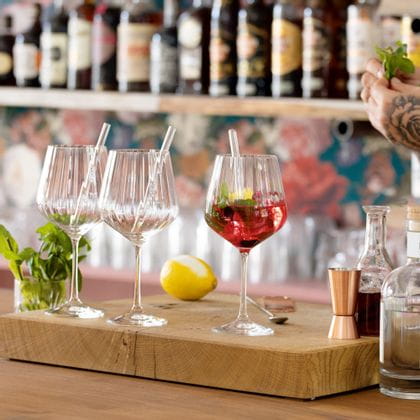 NACHTMANN Gin and Tonic glasses with glass straws on a bar counter. One is filled with a red Gin Cocktail with mint and a lemon zest. Behind the glasses is a lemon and a glass with mint sprigs. On the right are the hands of the bartender and bar equipment.<br/>
