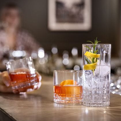 A bar counter with two SPIEGELAU Perfect Serve Collection glasses. The SOF glass is filled with an orange colored drink and an orange zest, the longdrink glass is filled with clear cocktail drink, ice cubes, rosemary and a lemon zest. In the background a hand, holding another filled SOF glass.<br/>