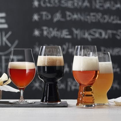 The with beer filled SPIEGELAU Craft Beer Glasses on a table in front of a blackboard with a menu. The glass for Barrel Aged beer and the Stout glass are standing on a slate plate, the IPA glass and the American Wheat Beer glass next to it on the table.<br/>