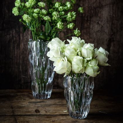 The small oval NACHTMANN Quartz vase filled with white roses in front of the tall NACHTMANN Quartz vase filled with white flowers on a sideboard made of dark wood.<br/>