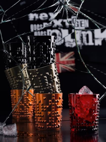 Four NACHTMANN Punk tumblers in jet-black, copper and gun metal stacked up in front of a broken mirror. Besides them the Punk tumbler in ruby filled with ice cubes. In the mirror there is a Junion Jack and the line nextgen goes punk.<br/>